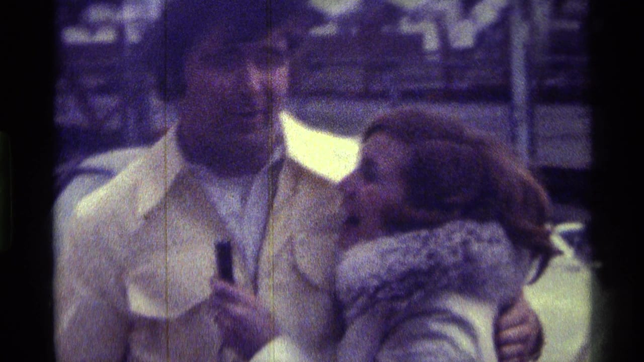 In 1976 Kentucky home-movie footage, a man in a light jacket stands beside a woman in a fur-trimmed coat near parked cars.