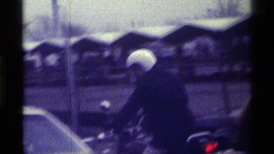 A blurred 1970s Kentucky street or parking-area view shows a person near a car with several parked vehicles in the background.