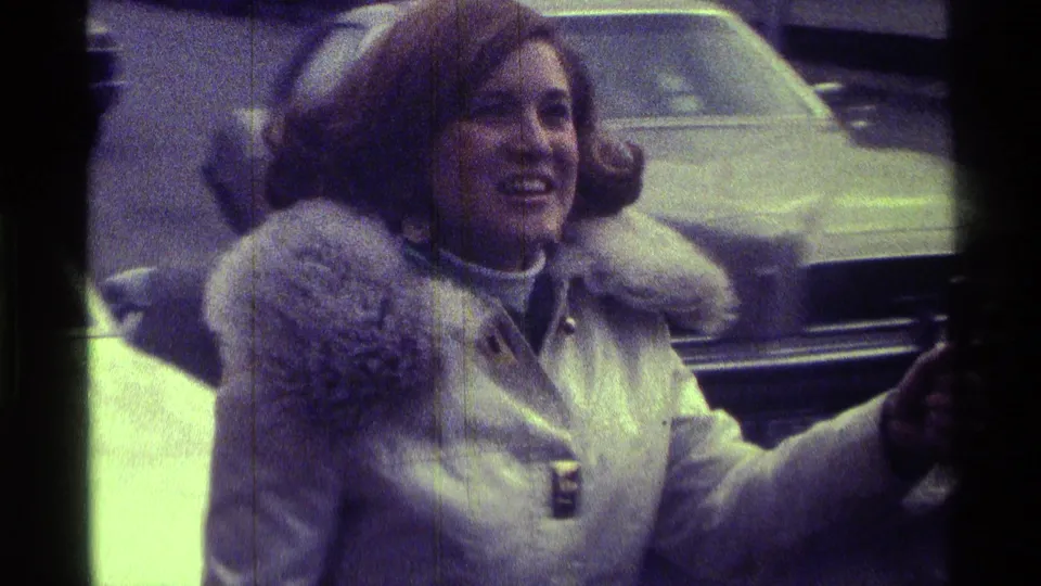 A smiling woman in a fur-trimmed coat stands beside a light-colored car in 1976 Kentucky home-movie footage.