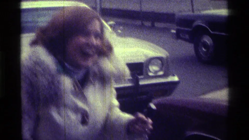 In a soft 1976 Kentucky frame, a woman in a fur-trimmed coat moves near the front of a parked car with another car behind.