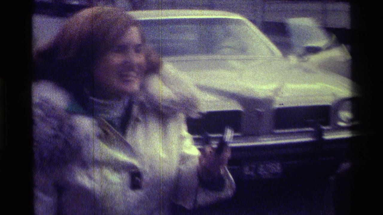 Smiling woman in a fur-collared coat stands near the front of a parked car.