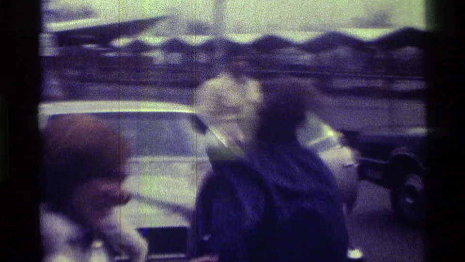 In the 1976 Kentucky home-movie frame, adults appear to confront or restrain one another beside parked cars in a public outdoor area.