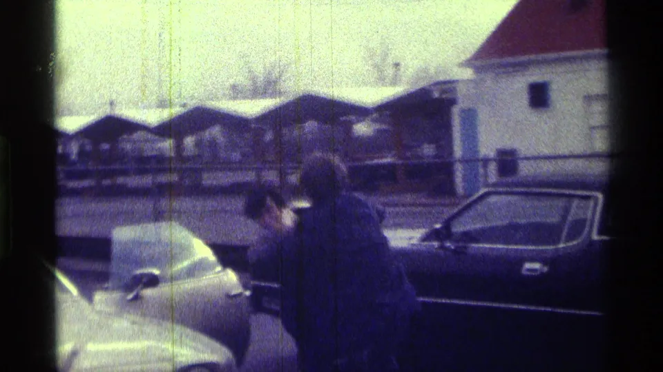 The 1976 Kentucky frame shows adults by classic cars, with a fence and low buildings behind them in a roadside or parking-lot setting.
