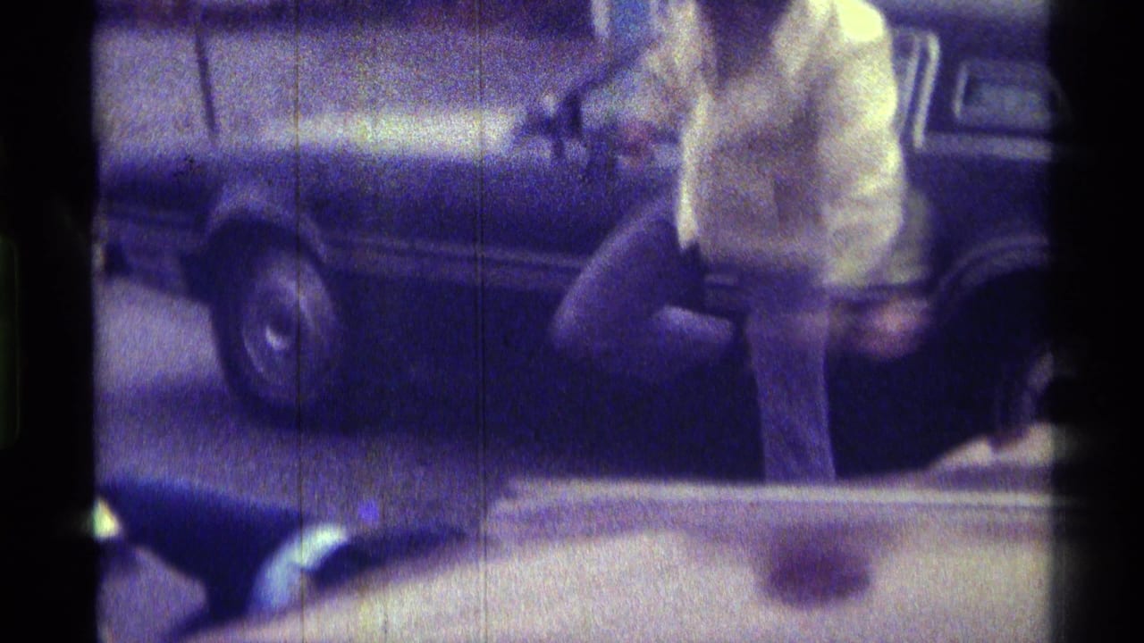Man in a light shirt bends forward beside a dark parked car with another person low in frame.