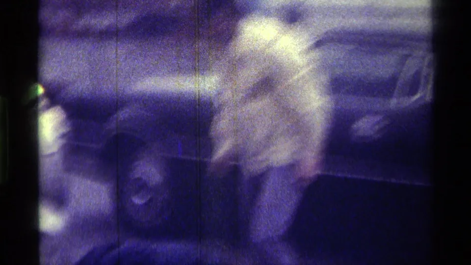 The handheld 8mm frame shows the man near the side of a dark car, with another figure partly visible at the edge.