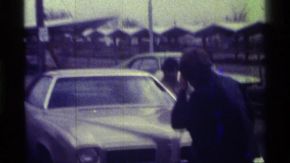A man in dark clothing bends beside a white car in a Kentucky parking lot, with another person partly visible at the edge of frame.