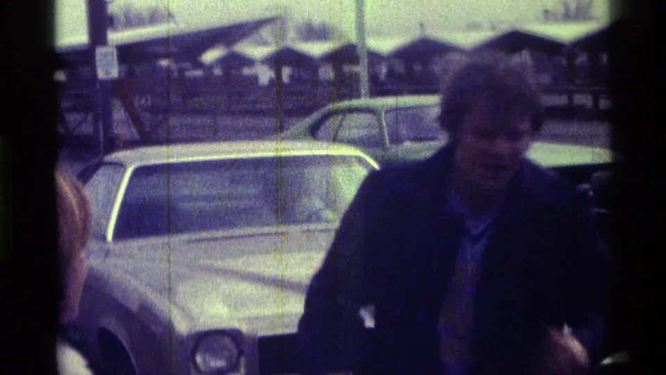 A man in a dark jacket and light shirt faces the camera near a white car, with a covered structure and parked cars behind him.