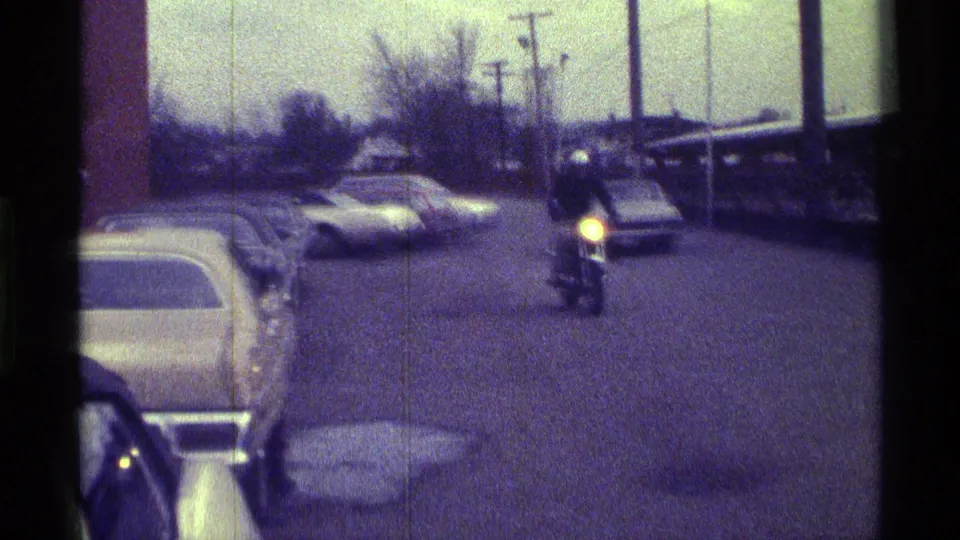 A motorcyclist rides across the parking lot in 1976 Kentucky, passing parked cars and utility poles under daylight.