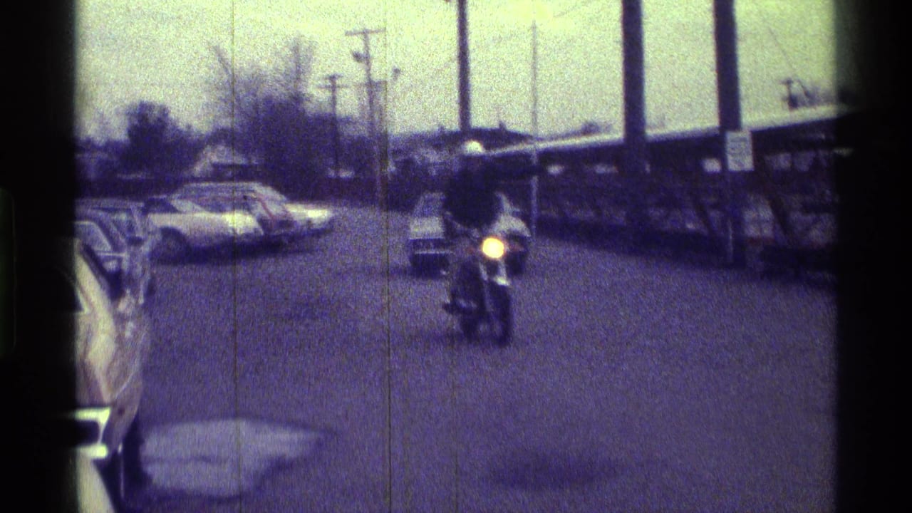 Motorcycle rider approaches on a quiet street with parked cars and utility poles.