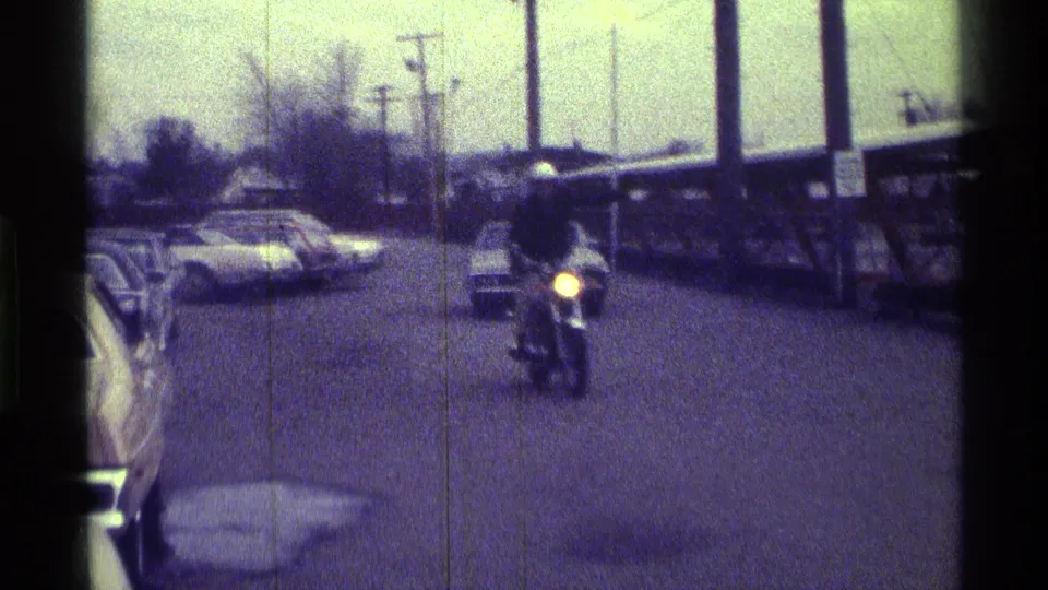 In Kentucky 1976 home-movie footage, a helmeted motorcycle rider approaches along a street lined with parked cars and utility poles.