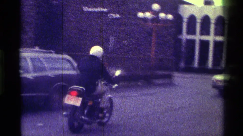 The rider continues away on the street near parked cars and a brick-looking building with tall arched window openings.