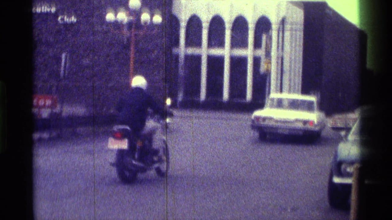 A motorcyclist rides through a Kentucky street scene in grainy 1970s 8mm footage, with older cars and building fronts visible.