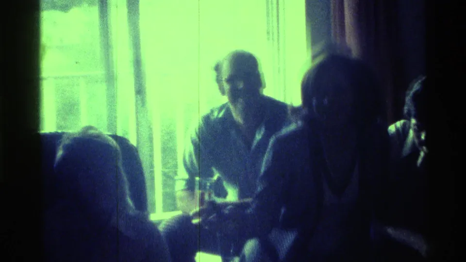 Adults sit together in a dim Kentucky interior, backlit by a bright greenish window in 1970s home-movie footage.