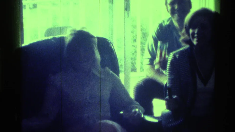 Several adults sit close together in a backlit private interior, with casual clothing and soft 8mm grain visible.