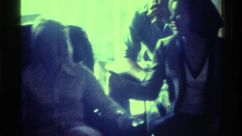 A woman appears to laugh while seated with friends in a dim 1970s Kentucky interior, rendered in green-blue 8mm exposure.
