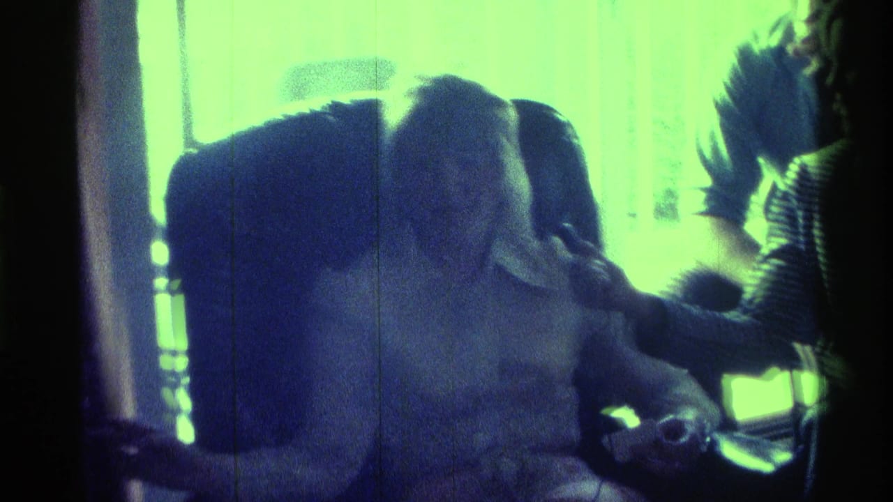 1976 Kentucky home-movie frame showing a seated person in a dark armchair with a microphone held near their face by another person.