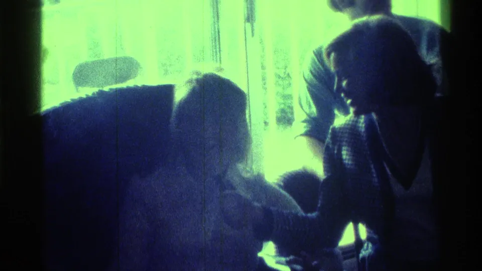 1976 Kentucky home-movie frame with a standing person leaning over a seated guest near chairs and a strongly backlit window.