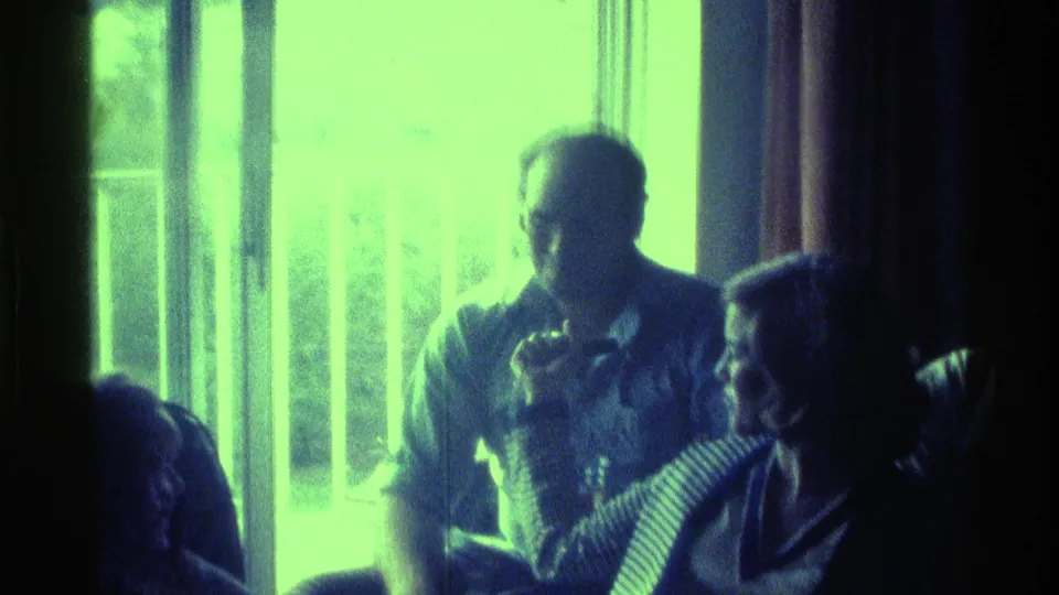 1976 Kentucky home-movie frame showing adults seated near a bright curtained window during an indoor birthday gathering.