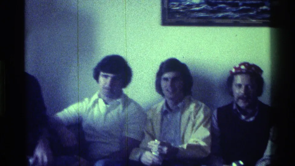 1976 Kentucky home-movie frame of three young men seated against a wall, one wearing a patterned cap.