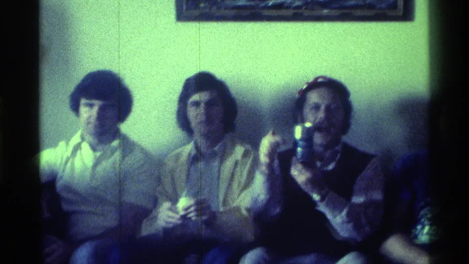 1976 Kentucky 8mm frame showing friends seated indoors as one man in a cap drinks from a small container.