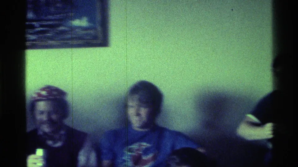 1976 Kentucky home-movie frame with two men seated near a pale wall and a standing figure partly visible at right.