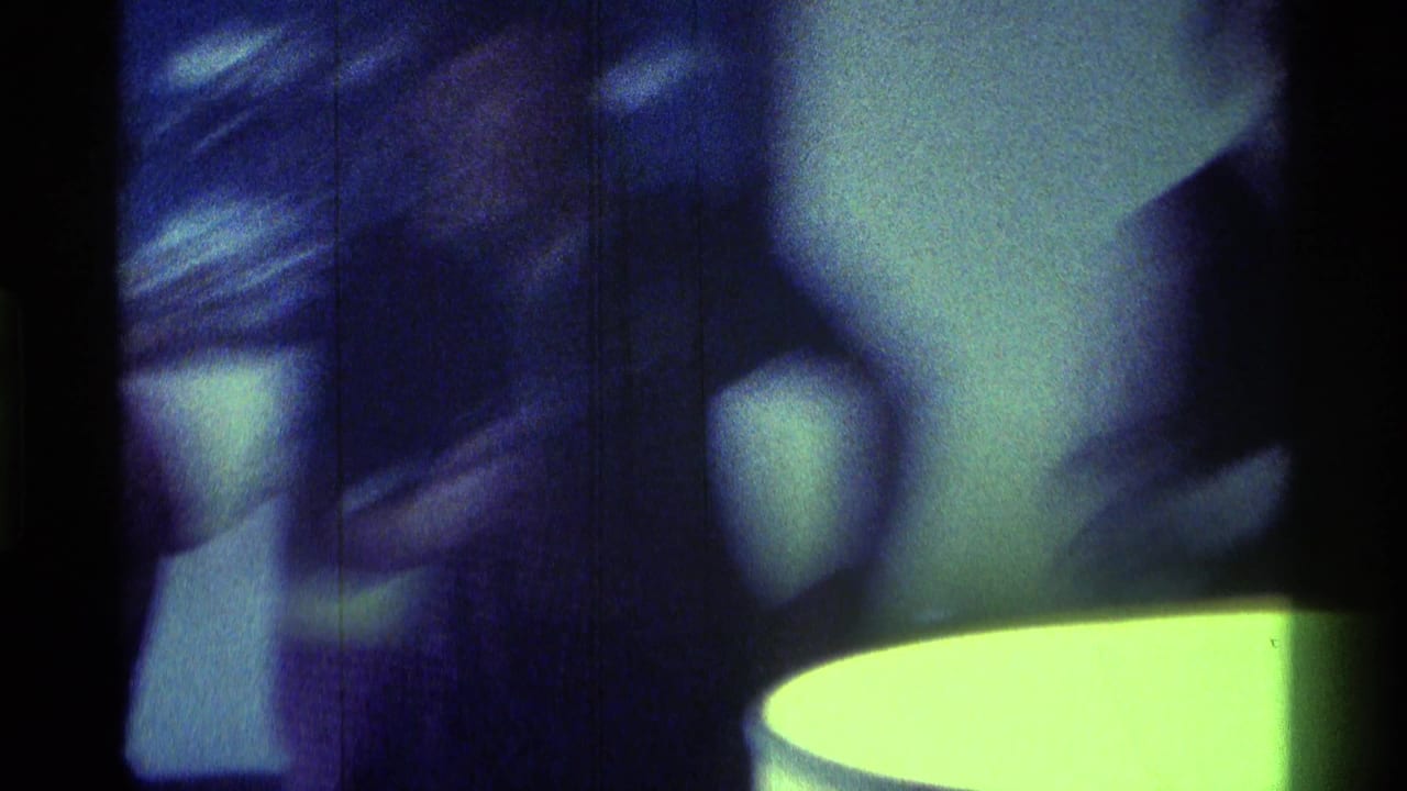 1976 Kentucky 8mm frame showing adult men in a dim private room, with soft focus, blue tint, and a bright round light or table edge in the foreground.