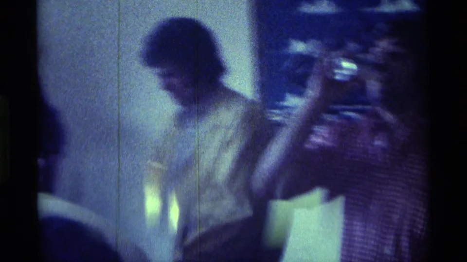 1976 Kentucky home-movie frame with two adult men seated close in a dim interior, their faces softened by grain, low light, and blue color shift.