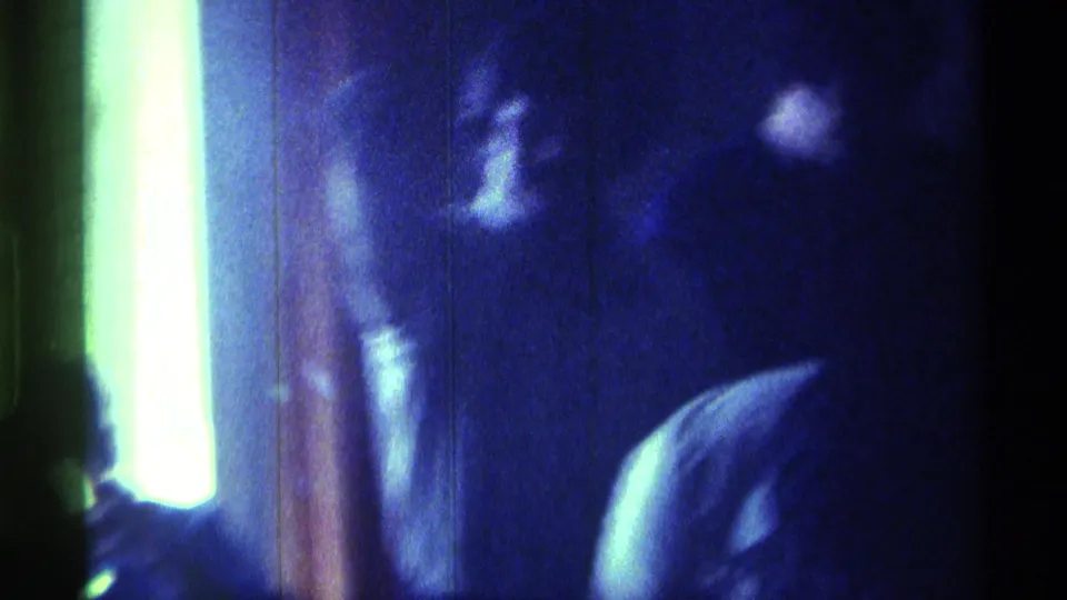 Blue-tinted 8mm frame from a private Kentucky interior, showing a bright vertical light source and indistinct adult figures in soft handheld blur.