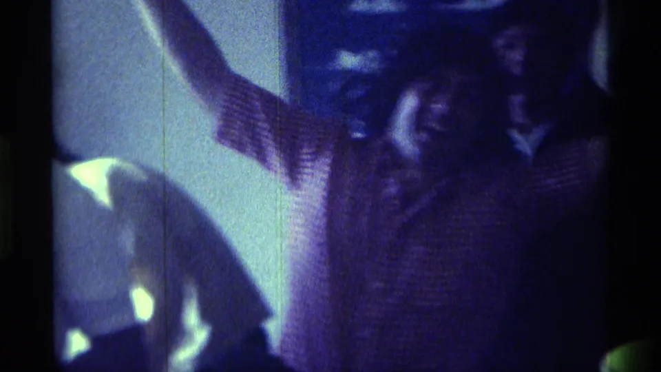 1976 Kentucky 8mm frame of an adult man in a striped shirt raising his arms in a dance-like gesture inside a dim private room.