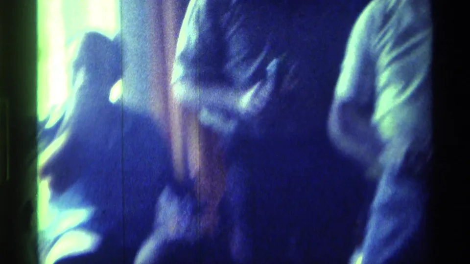 Blurred 8mm frame of adults in a private room, with raised arms and close bodies suggesting dancing or celebration.