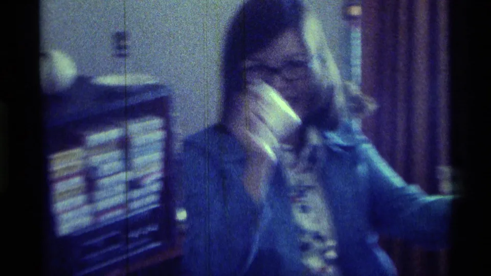 1976 Kentucky home-movie frame of a smiling woman in denim holding a stack of cash inside a private room.