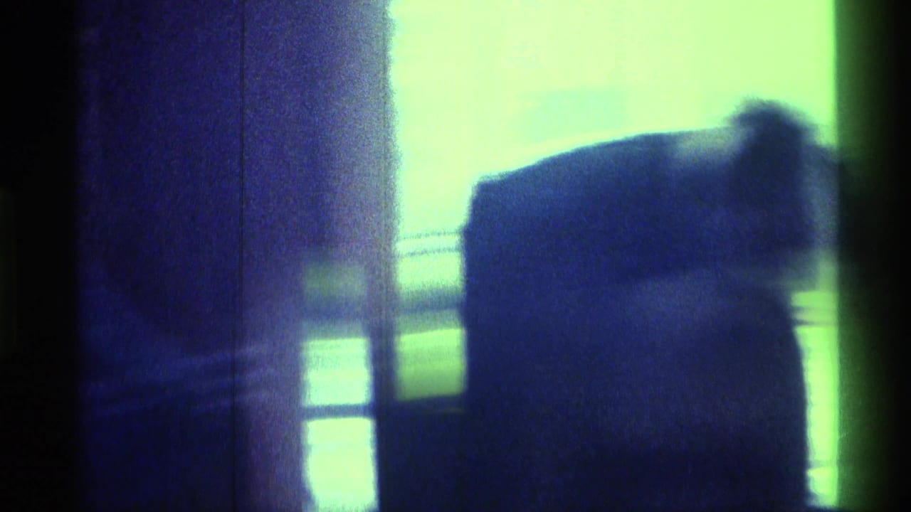 Dark interior frame with a seated figure silhouetted near a bright window or wall.