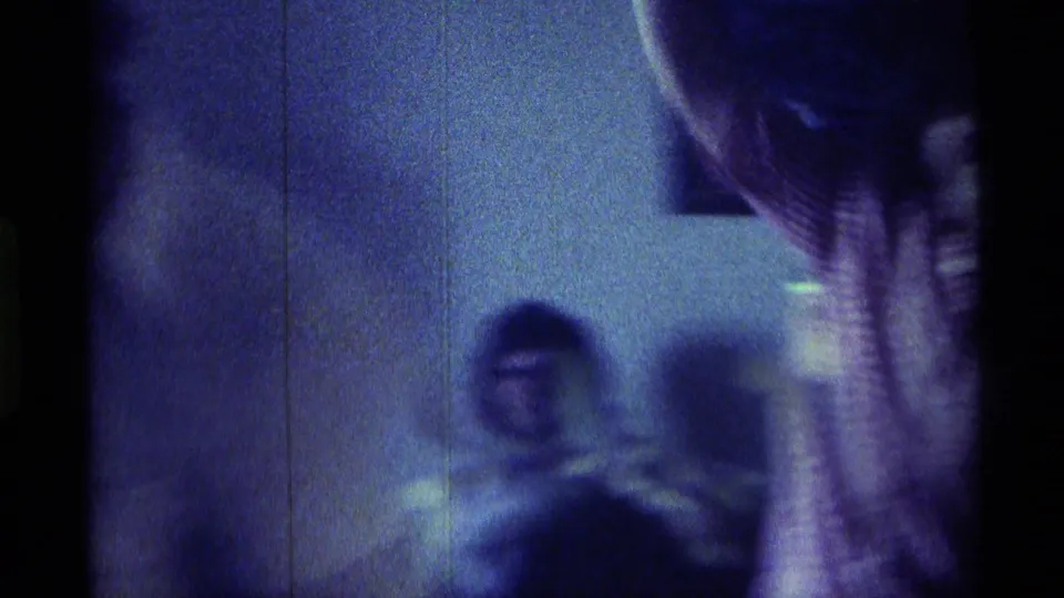 1976 Kentucky interior frame with a blurred seated adult against a wall and a partial person near the right edge in handheld 8mm footage.