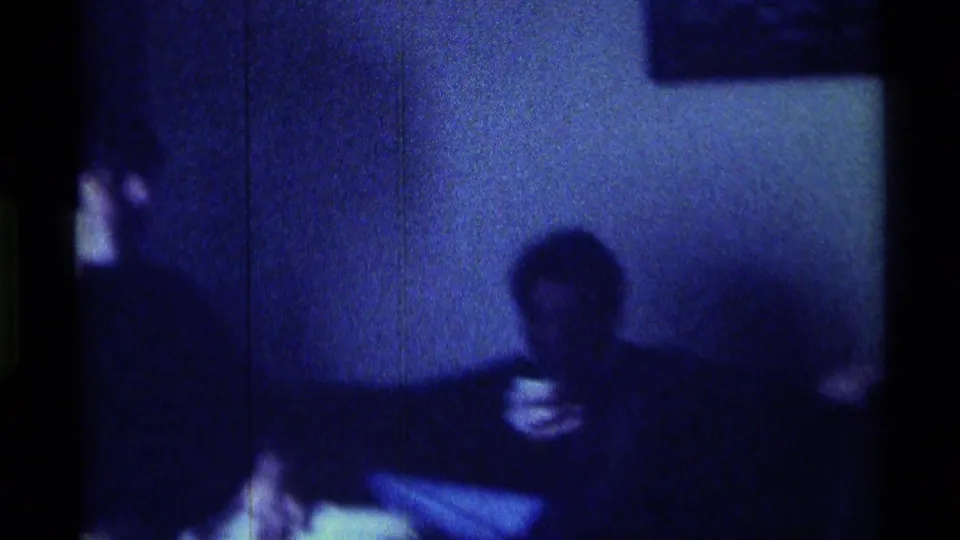 1976 Kentucky home-movie frame showing a shadowed adult seated on a sofa or low seat, apparently holding a cup during an indoor gathering.