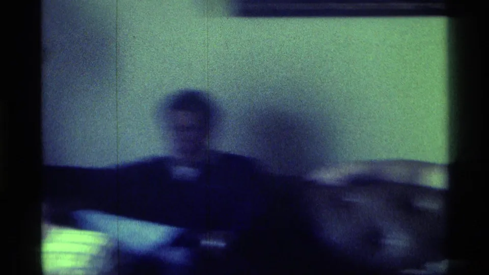 1976 Kentucky indoor frame showing a seated adult with arms spread across low seating, surrounded by soft focus, shadows, and green-blue film color.