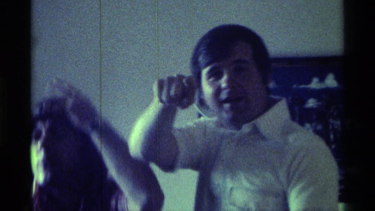 Two people indoors, with a young man in a white shirt gesturing toward the camera.