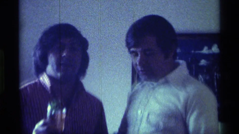 1976 Kentucky 8mm frame of two young men indoors, one in a striped shirt holding a drink-like object and one in a white shirt.