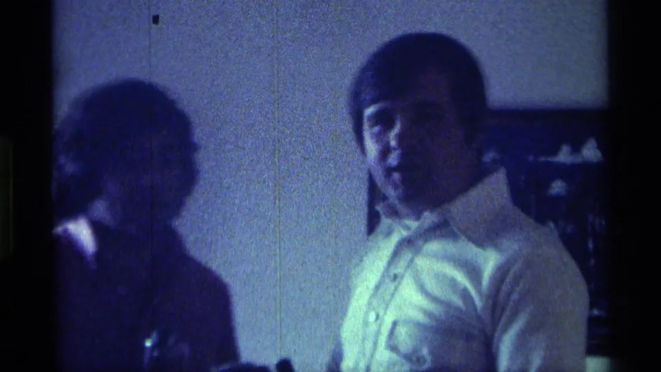 1976 Kentucky 8mm frame with a young man in a white collared shirt facing the camera beside another person in a dim room.