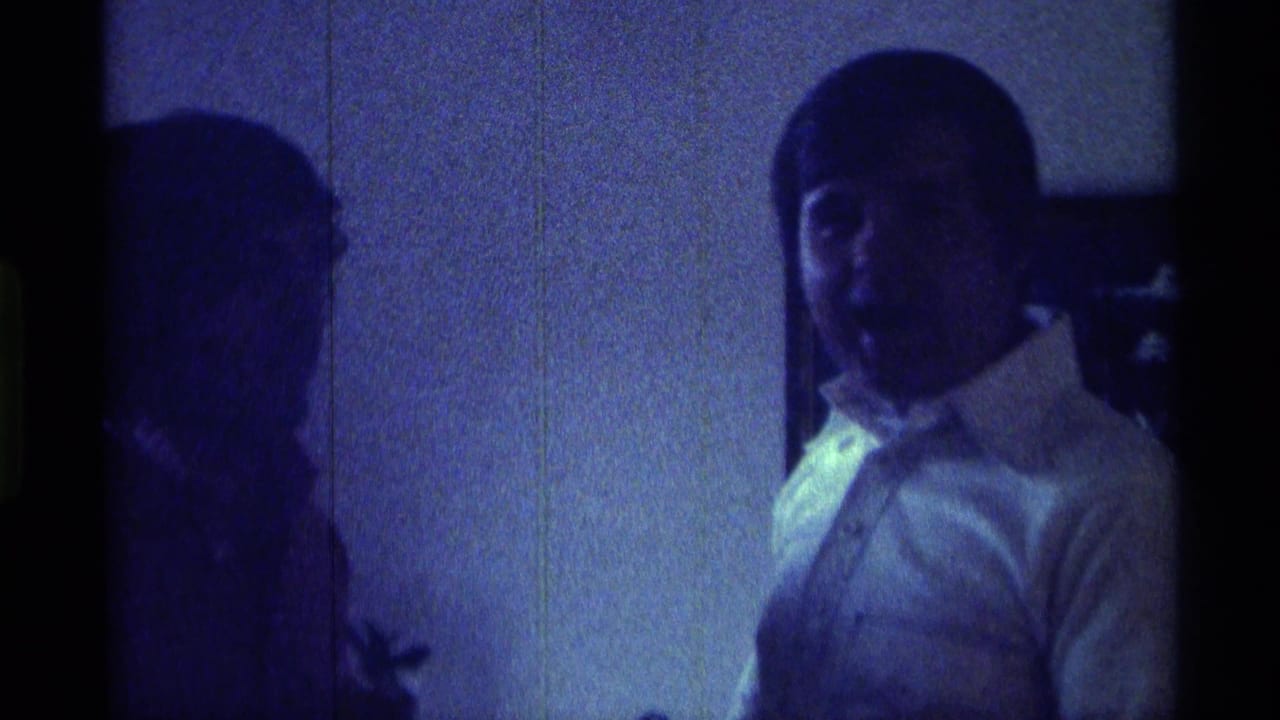 Man in a white collared shirt faces the camera beside a shadowed person indoors.