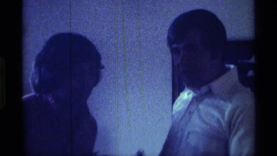 The man remains at the right side of the blue-tinted indoor frame, with a second person partly visible in silhouette at left.