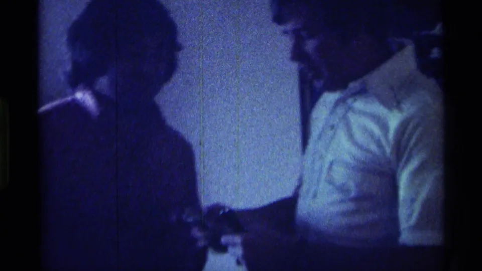 In the dim 8mm interior, the man looks down toward a small object in his hand while another person stands close in shadow.