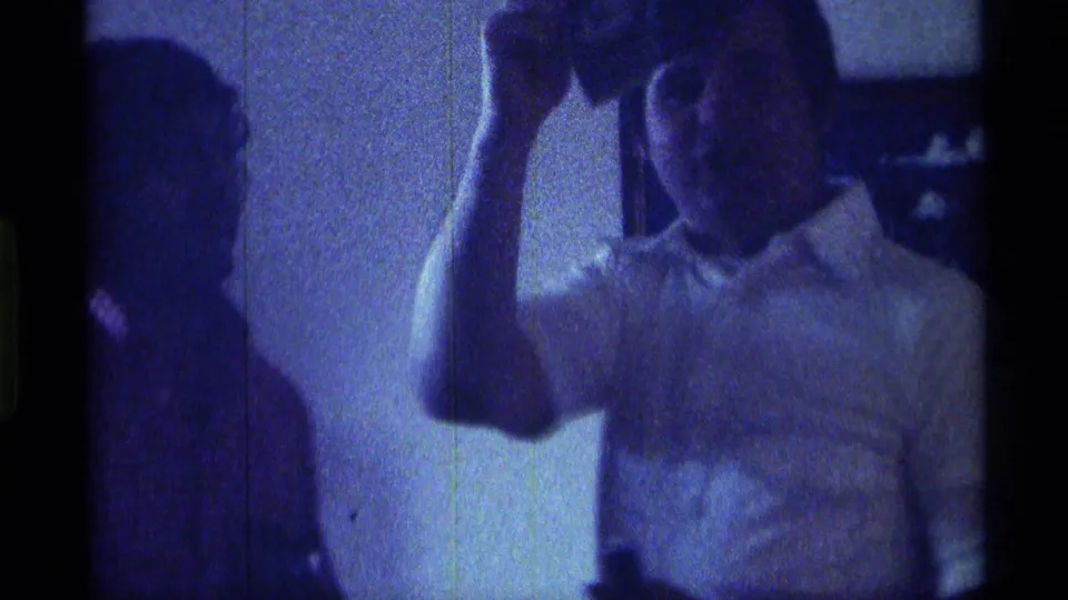 The man lifts a small object near his head and faces the camera, with another person faintly visible beside him in the 1976 Kentucky interior.