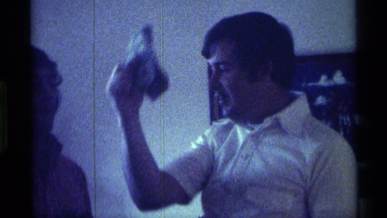 In a dim 1976 Kentucky interior, a young man in a light collared shirt raises a small object while speaking to another man.