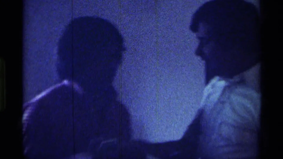 Two young men stand close together in a dim Kentucky room, with one figure mostly in shadow and the other in profile.