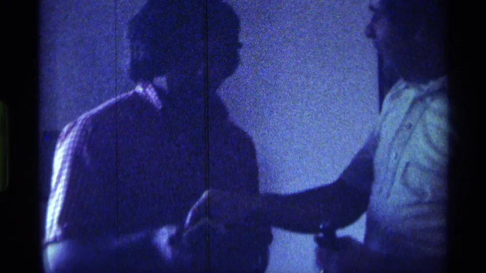 A young man in a striped shirt faces a man in a light collared shirt during a close indoor exchange in 1976 Kentucky footage.