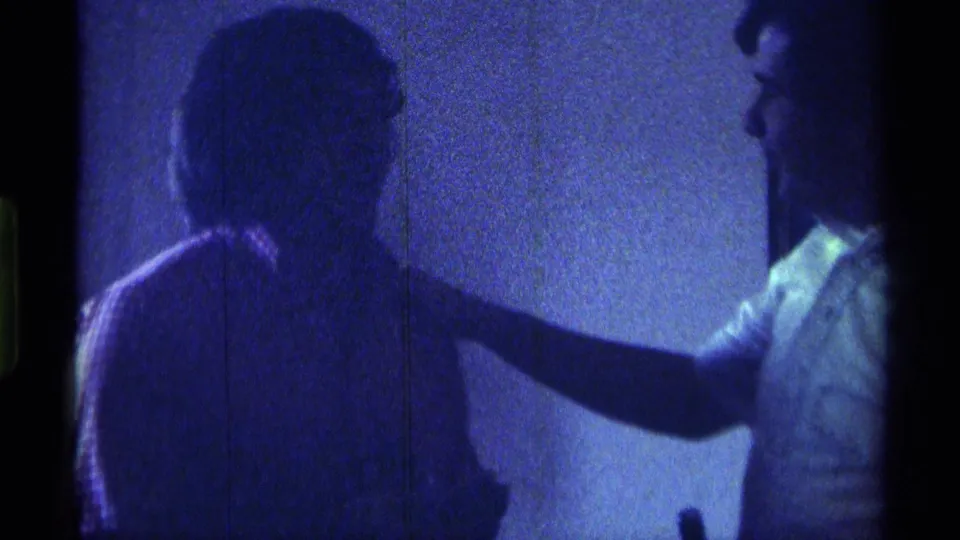 In a blue-tinted indoor frame, a man in a light shirt extends his arm toward another young man whose face is in shadow.