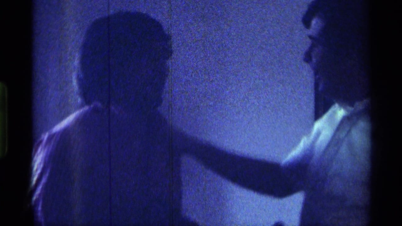 Two adult men in a dim blue-lit room, one reaching toward the other's shoulder.