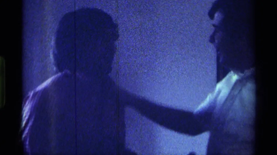 1976 Kentucky 8mm frame of two adult men indoors, with one man reaching toward the other's shoulder in dim blue light.