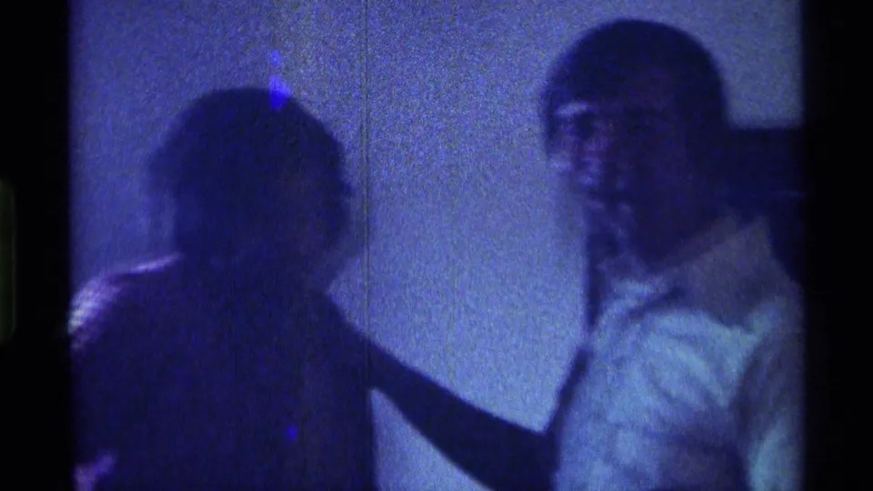 1976 Kentucky home-movie frame with two adult men in a blue-green interior, one partly turned toward the camera.