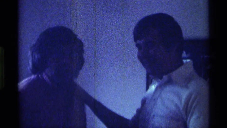 1976 Kentucky 8mm frame of two adult men in a dim interior, with one turned in profile beside a shadowed figure.